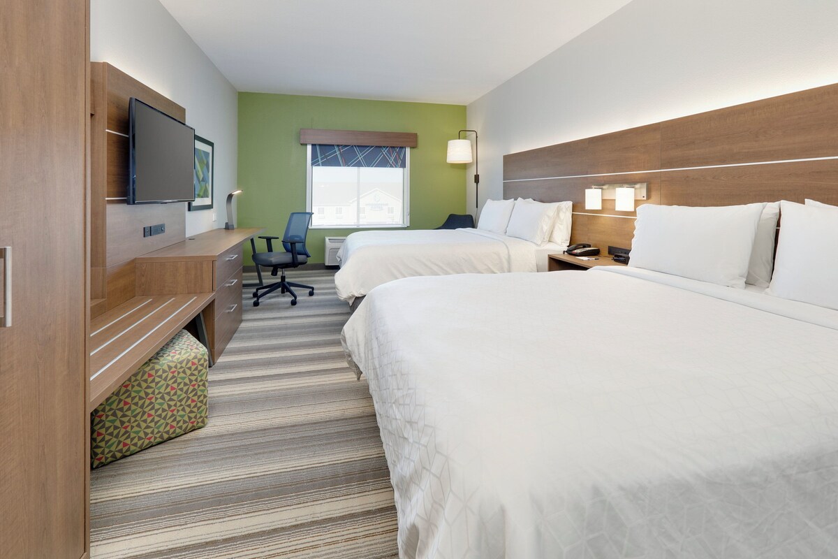 A hotel room with two beds, a desk, a chair, a TV, and a window with a view of a building.