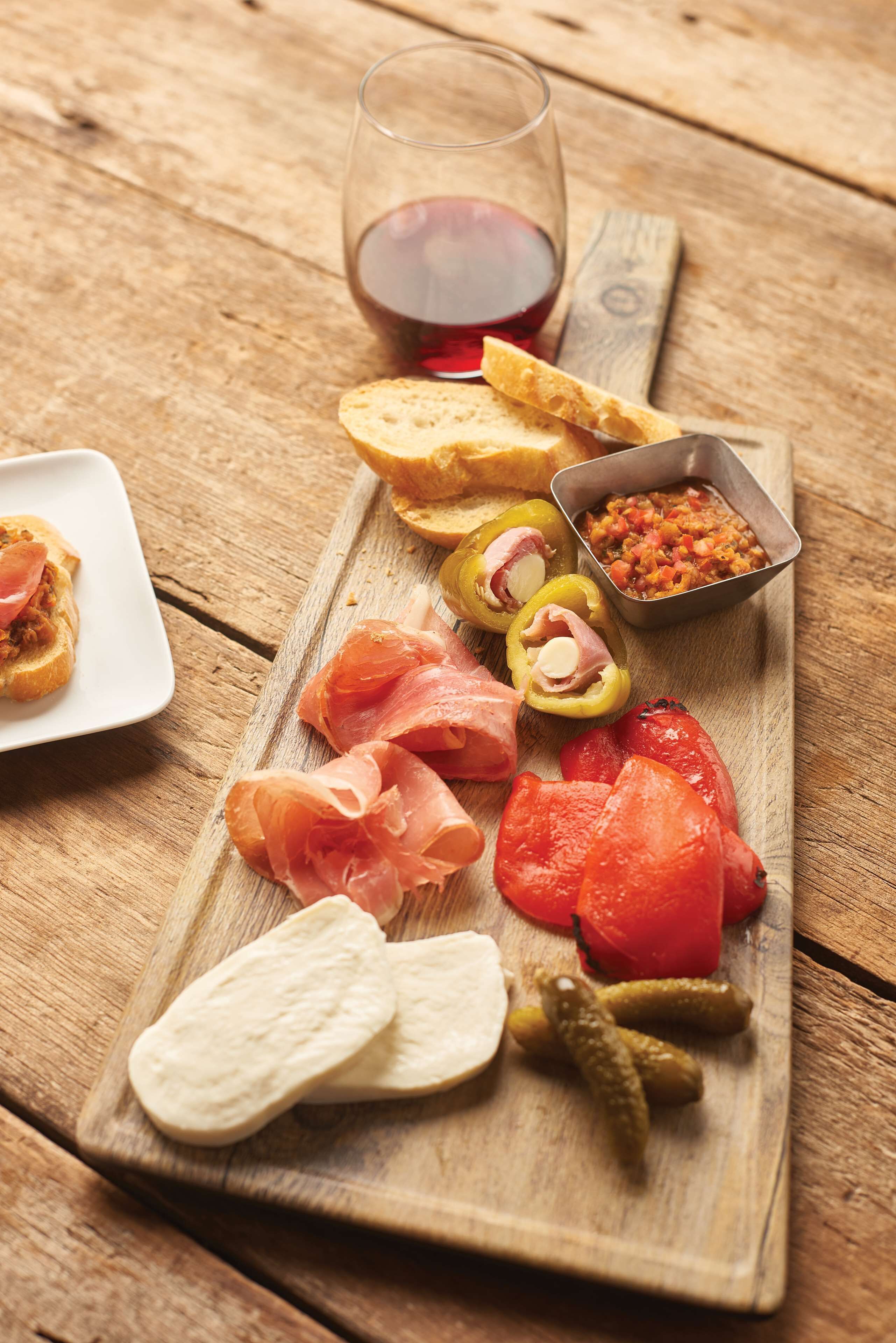 A wooden cutting board with various food items including bread, tomatoes, pickles, and a small bowl of salsa.