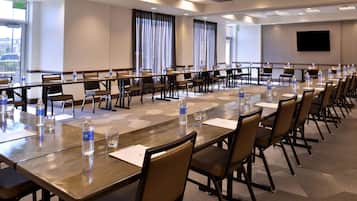 Una sala de conferencias con varias mesas y sillas, equipada con botellas de agua y documentos, y una pantalla grande en la pared.