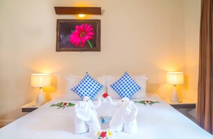A hotel room with a bed, two nightstands, two lamps, checkered pillows, a framed picture of a flower, and towels shaped like elephants.