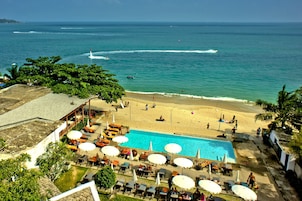 A pool area with lounge chairs and umbrellas, a beach with people, and a view of the ocean.