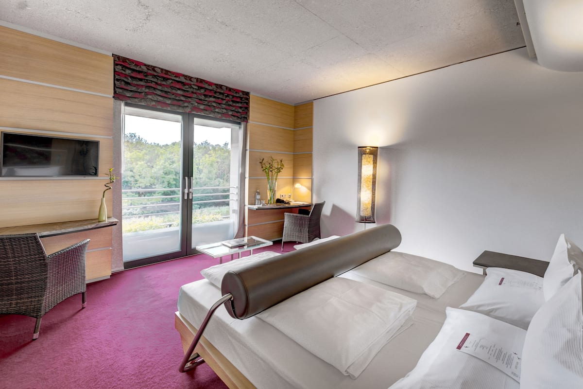 Quarto de hotel com cama, escrivaninha, cadeira, televisão, janela e varanda.