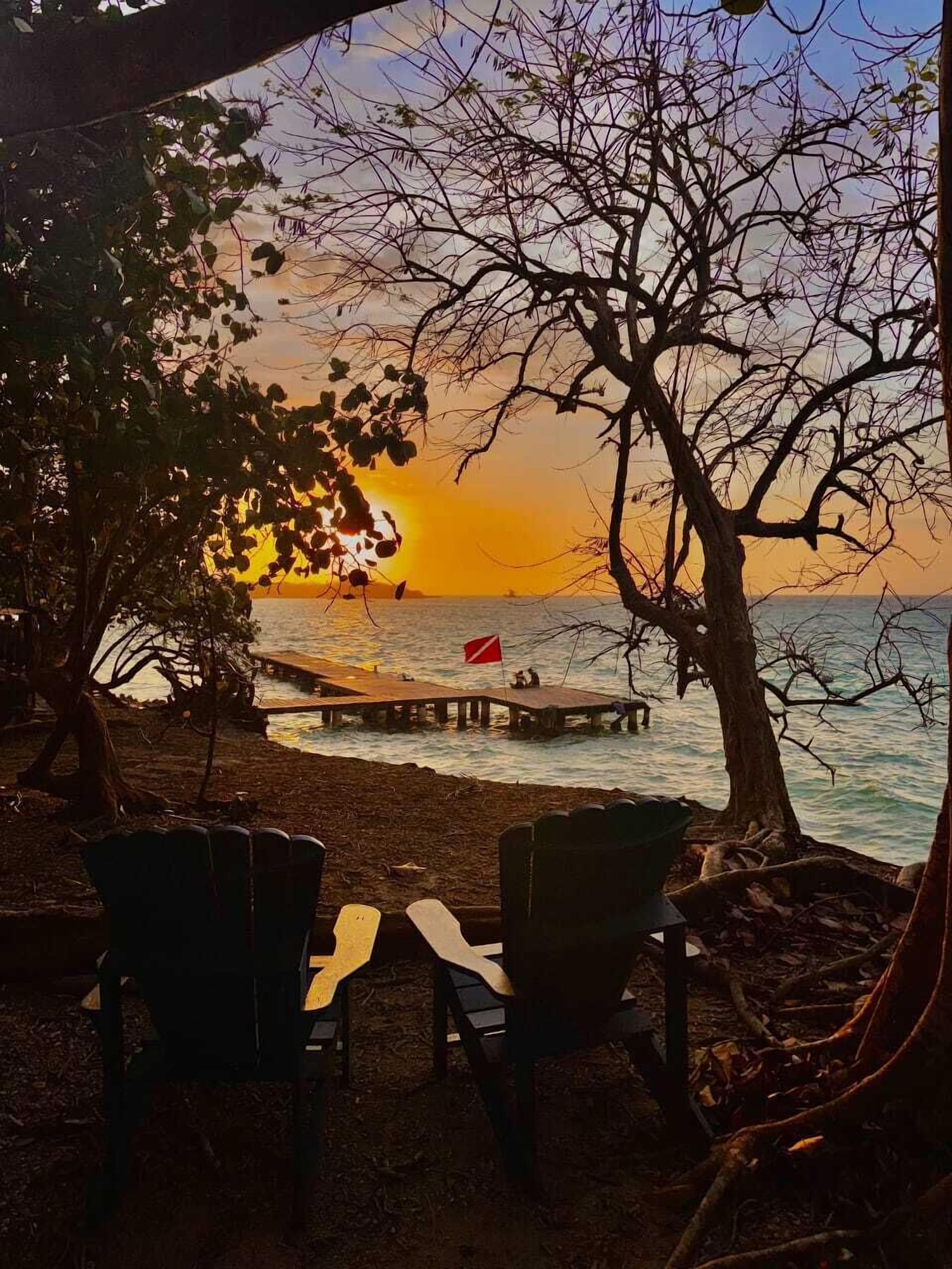 Un área de descanso junto a la playa con dos sillas y una mesa, con vista a la puesta de sol sobre el agua y un muelle al fondo.