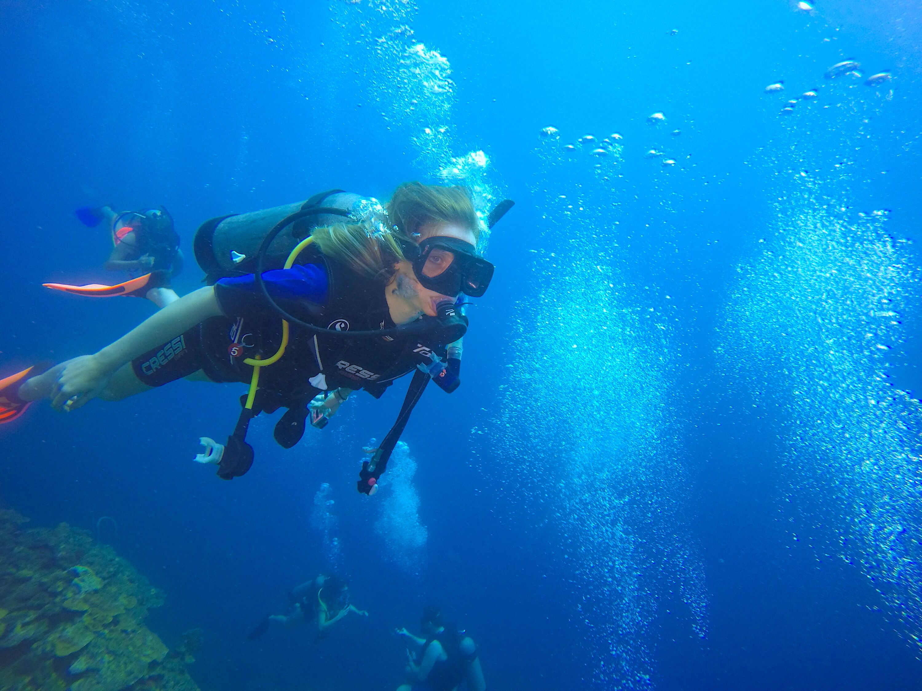 A diver underwater with a clear view of the ocean floor and other divers in the background.