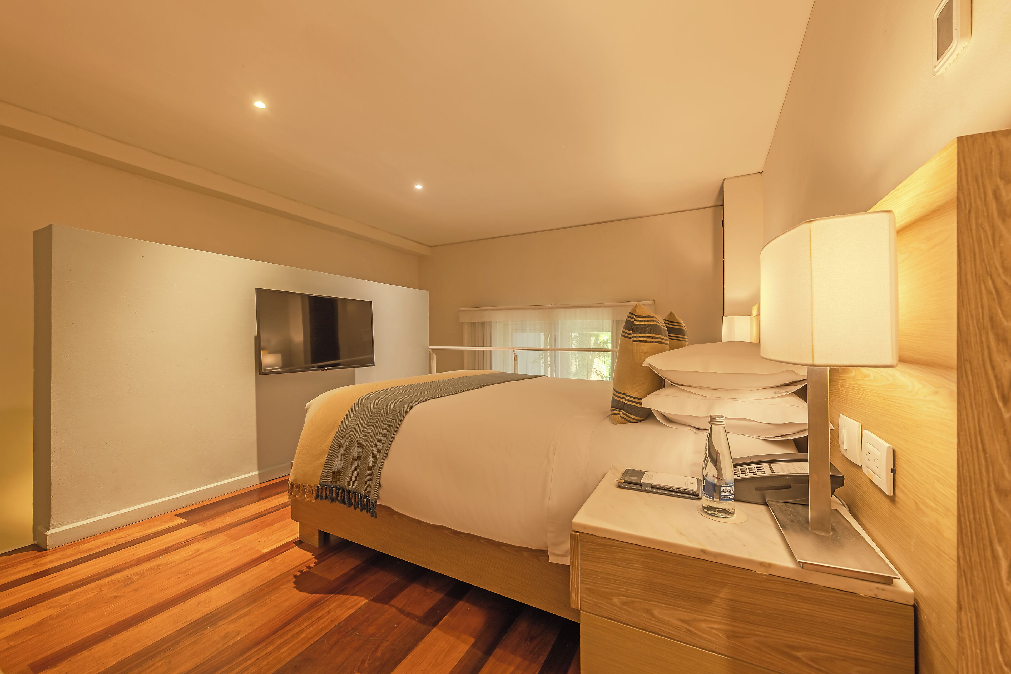A modern hotel room with a wooden floor, a large bed with a grey throw, a bedside table with a lamp, and a TV mounted on the wall.