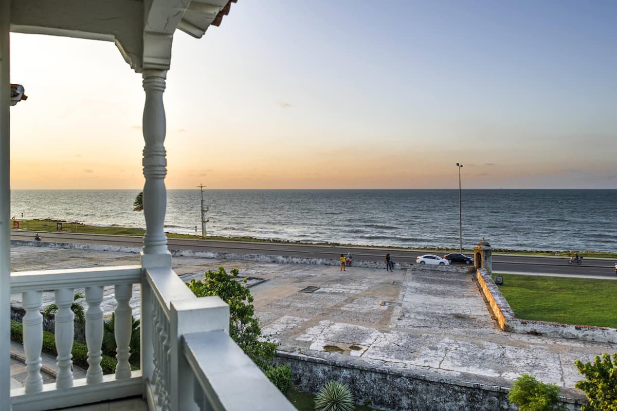 Blick auf die Küste vom Balkon aus, mit einem Steinweg, der zum Meer führt, einer Straße mit Fahrzeugen und einem klaren Himmel.