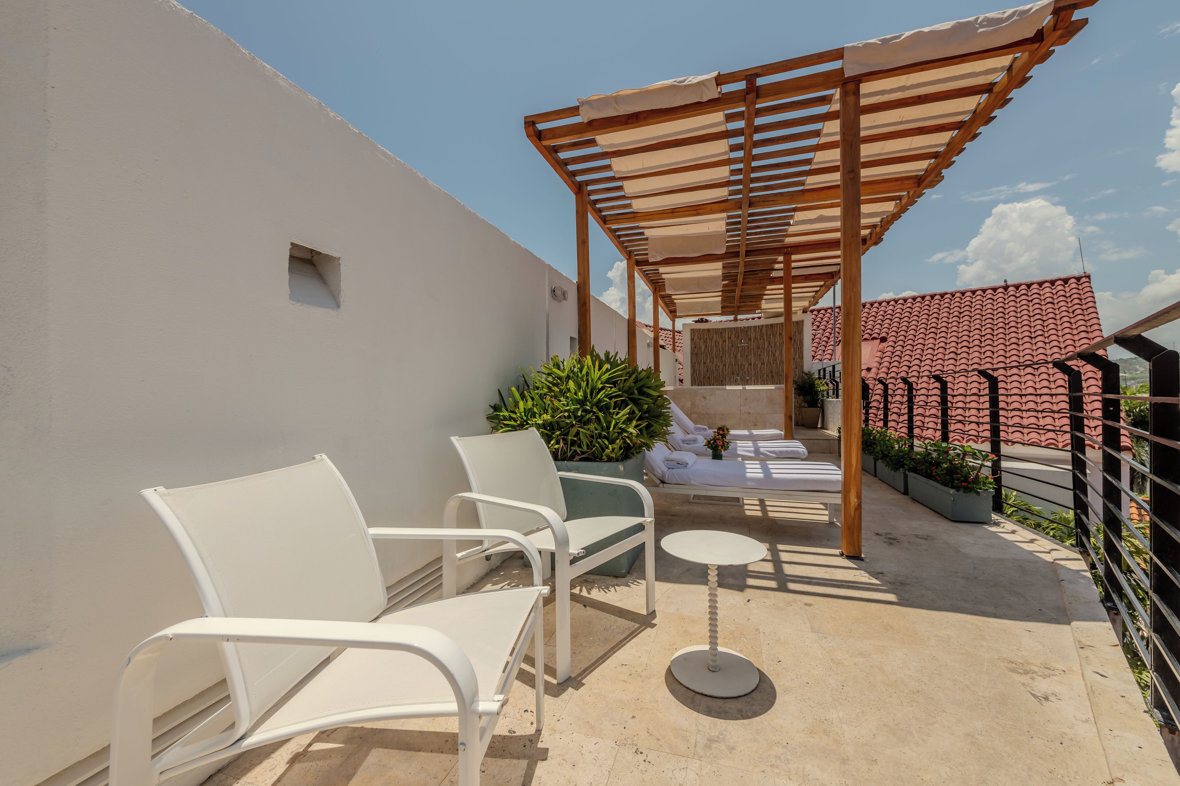Eine Dachterrasse mit weißen Metallstühlen, einem kleinen runden Tisch und einer Holzpergola, die Schatten spendet.