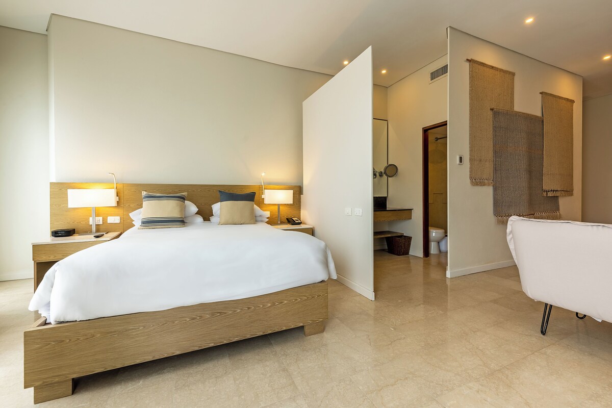 A modern hotel room with a wooden bed, bedside lamps, a white bathroom with a bathtub, and a mirror.