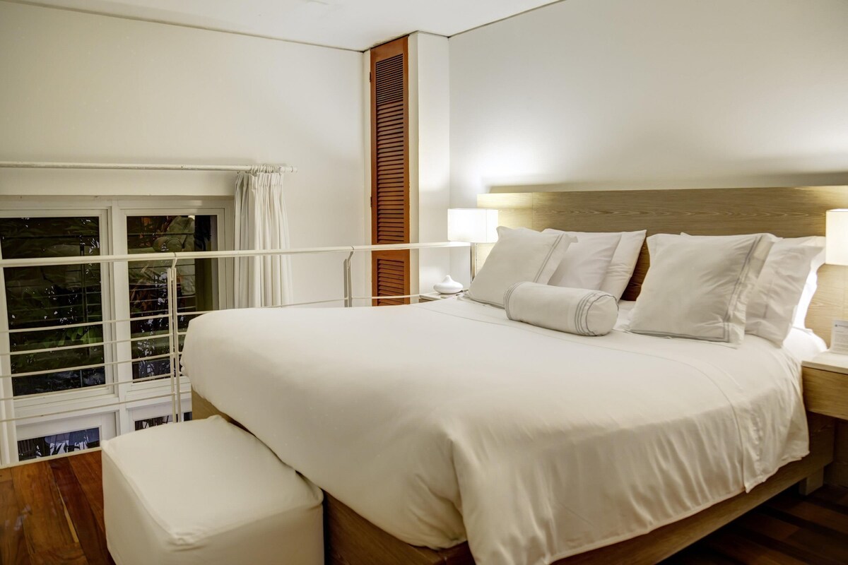 Un dormitorio con una cama grande, una mesita de noche, una ventana con persianas y vistas a la vegetación.