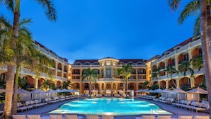 A hotel with a large pool surrounded by lounge chairs and umbrellas, with palm trees and a multi-story building in the background.