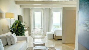 A modern hotel room with a white sofa, a small wooden coffee table, a metal chair, a bed, and a large potted plant.