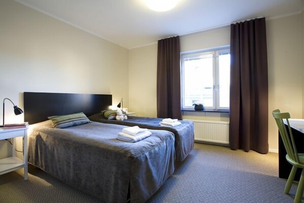 A hotel room with two beds, a desk with a lamp, a chair, and a window with curtains. - Hotel Bakfickan (Stockholm)
