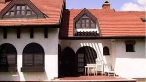 A two-story building with a red-tiled roof, white walls, and dark wooden trim. There are two visible windows with shutters, and a small outdoor seating area with a table and chairs.