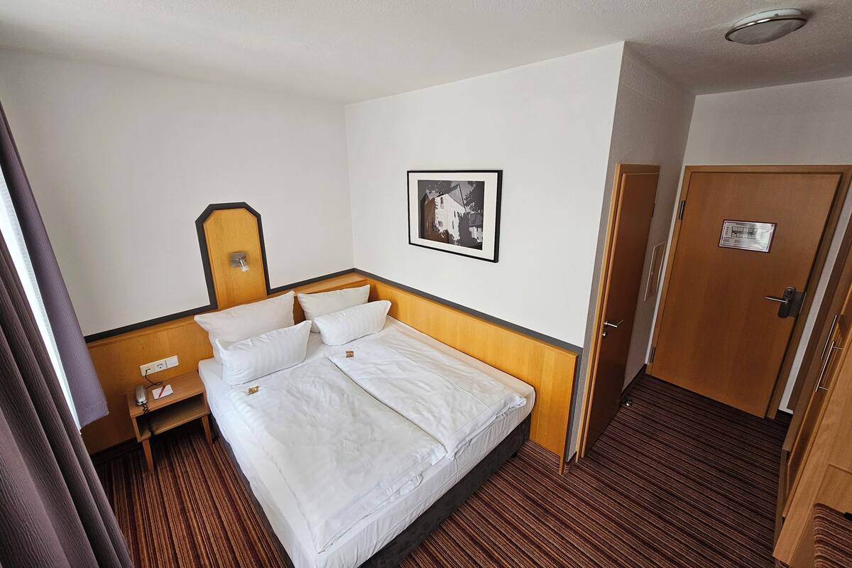 Ein Hotelzimmer mit Bett, Nachttisch mit Lampe, einem gerahmten Bild an der Wand und einer Tür mit Schild.