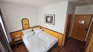 Habitación de hotel con cama, mesita de noche con lámpara, un cuadro en la pared y una puerta con un cartel.