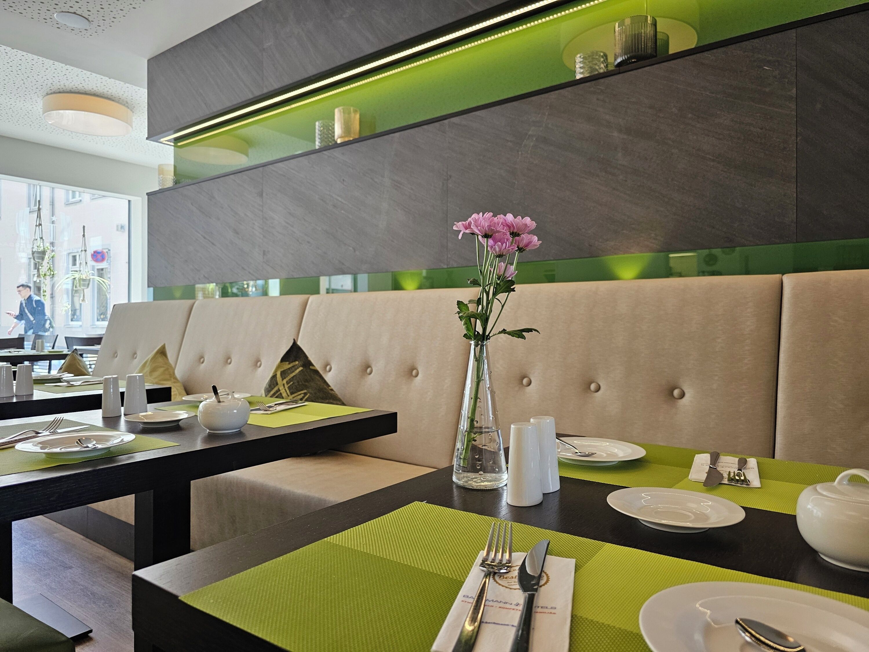 Un restaurante con una paleta de colores verde y beige, que presenta un área de asientos con bancos de respaldo alto, mesas bien dispuestas con platos blancos, y un jarrón con flores rosadas.