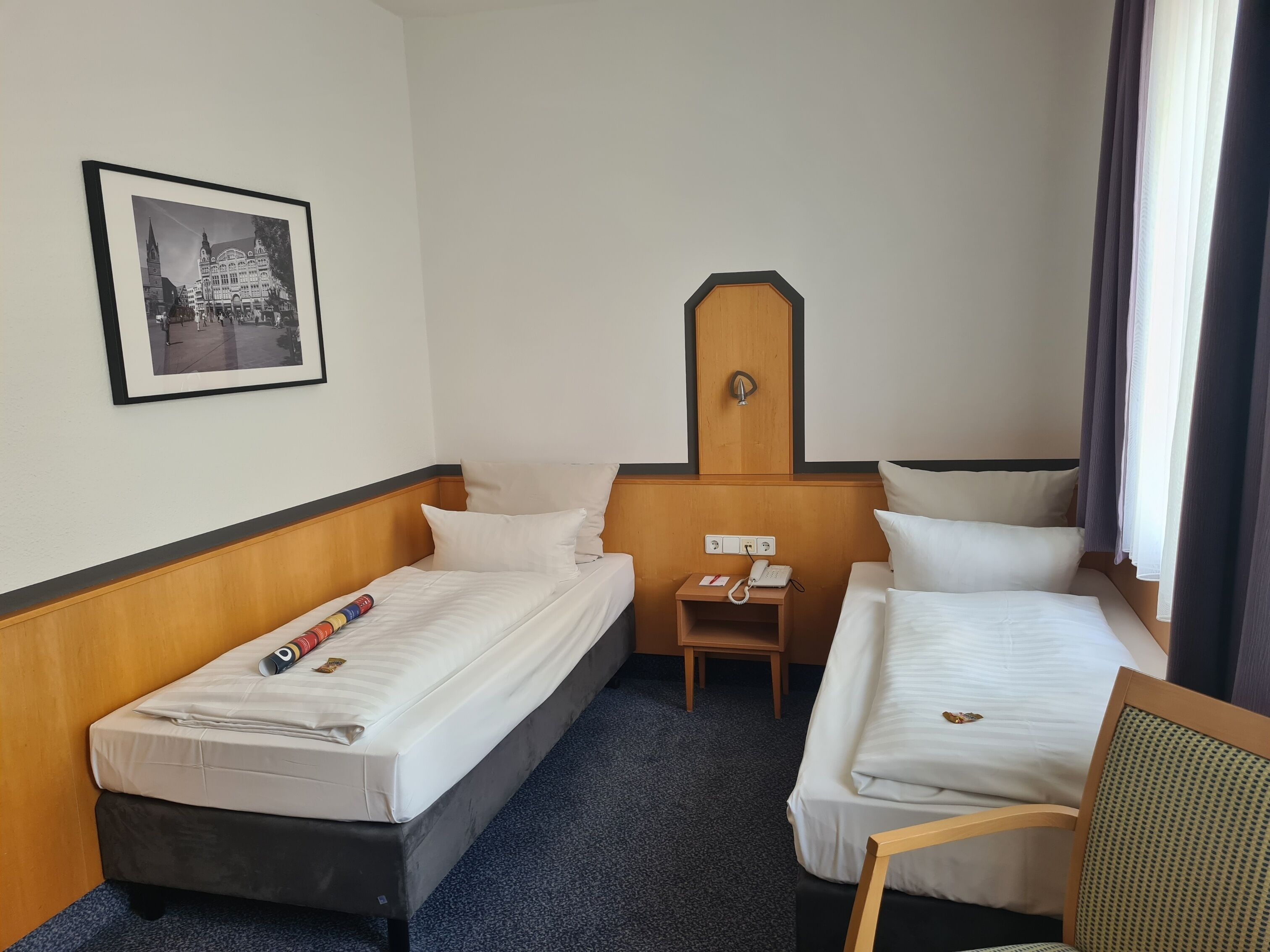 Una habitación de hotel con dos camas individuales, una mesita de noche de madera con un teléfono, un cuadro en la pared y una ventana con cortinas.