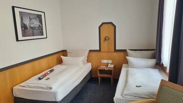 Una habitación de hotel con dos camas individuales, una mesita de noche de madera con un teléfono, un cuadro en la pared y una ventana con cortinas.
