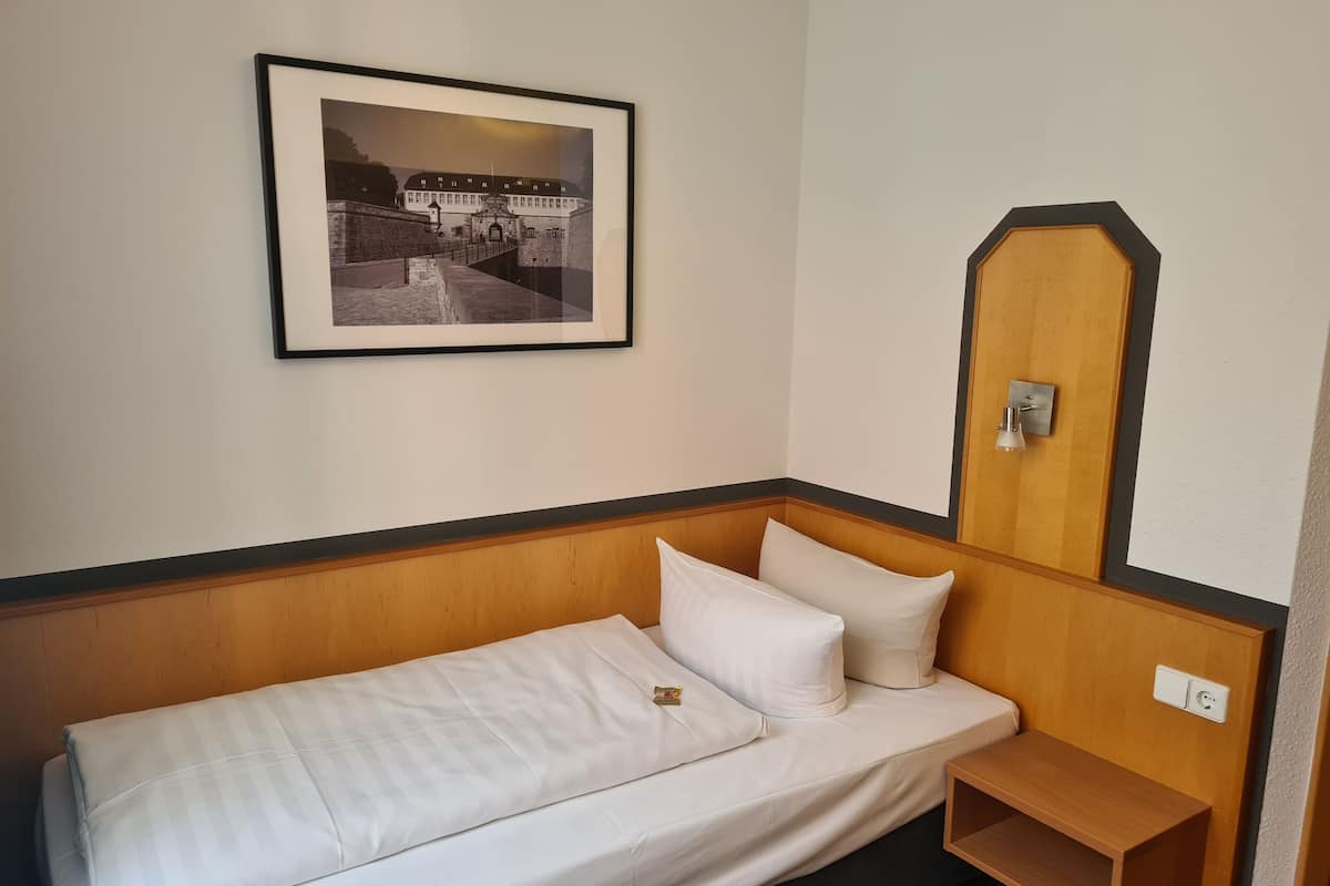 Ein Einzelbett mit weißer Bettwäsche, ein kleiner Nachttisch aus Holz, ein gerahmtes Bild an der Wand und eine Holztür mit Spiegel.