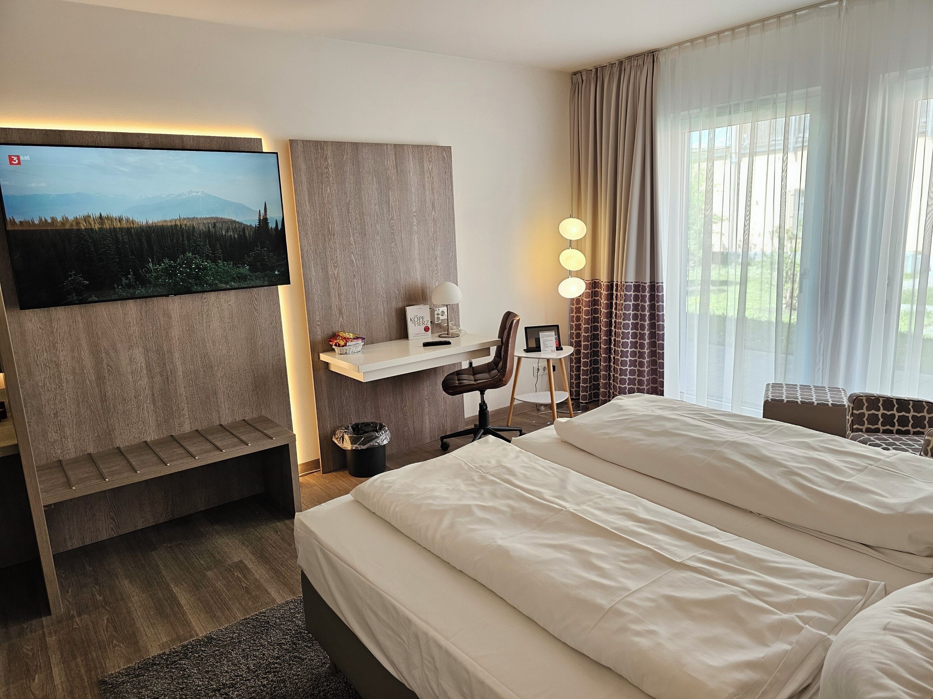 A hotel room with a bed, a desk with a chair, a TV mounted on the wall, and a window with curtains.
