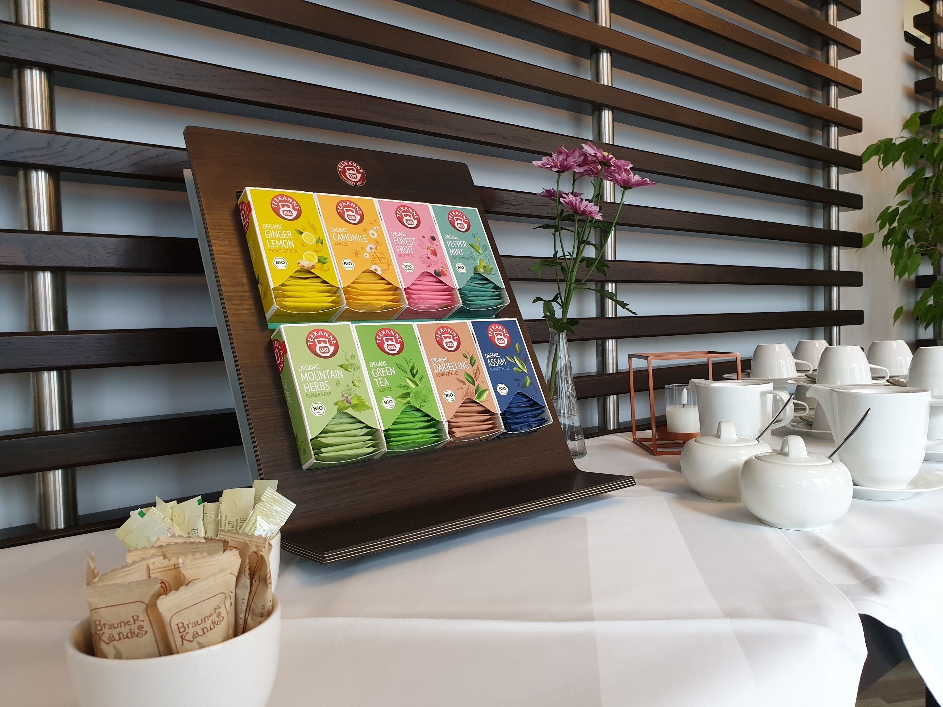 A tea display with various organic blends, a vase with flowers, and white tea sets on a table.