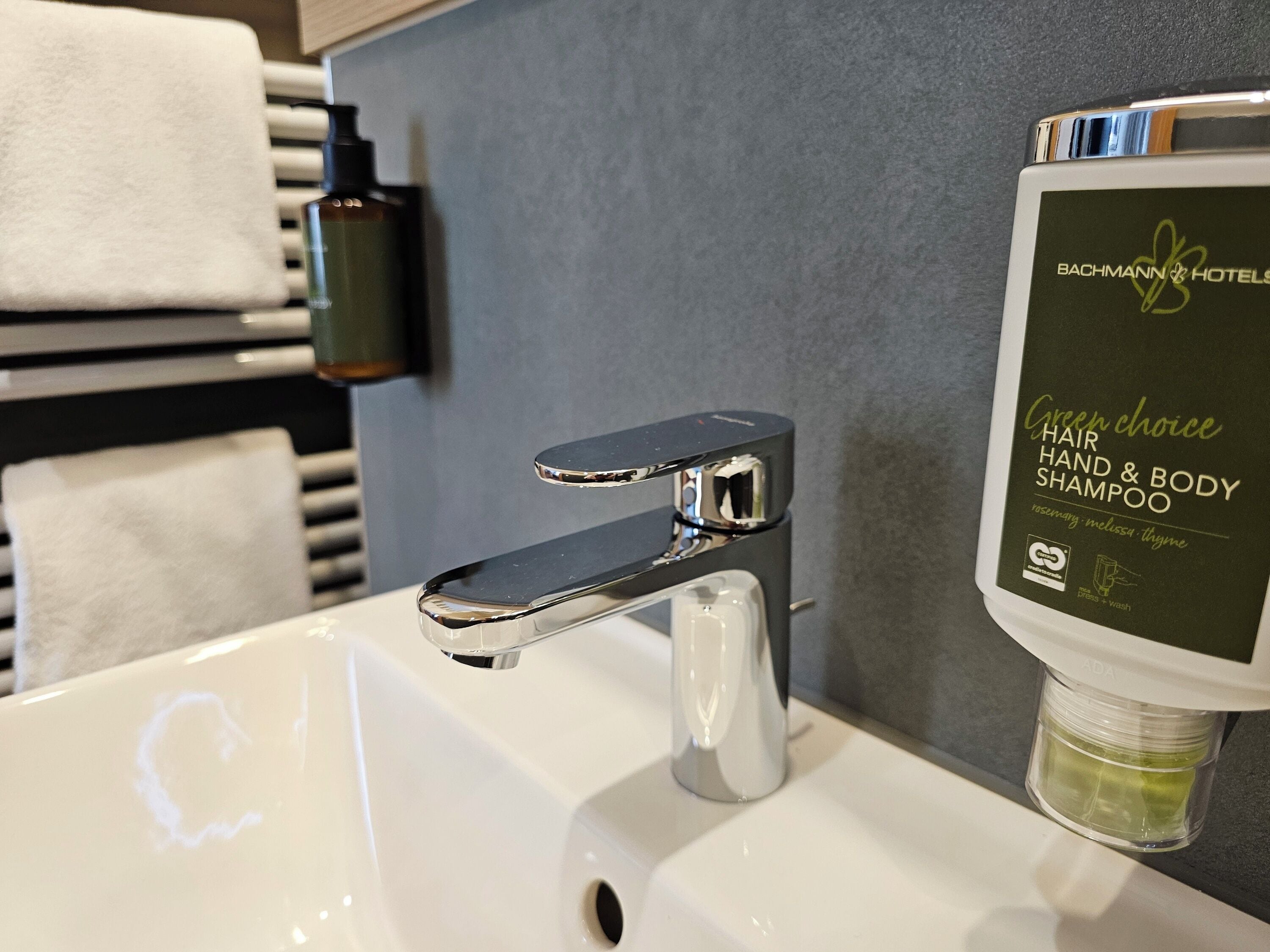 Un lavabo con rubinetteria cromata, una bottiglia di shampoo 'Green Choice' per capelli, mani e corpo, e un porta asciugamani con asciugamani bianchi.
