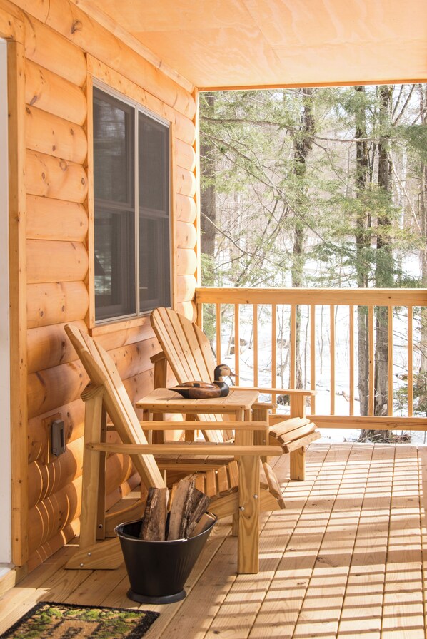 Une cabane en bois avec une véranda, deux fauteuils Adirondack, une table et un seau de bois de chauffage.