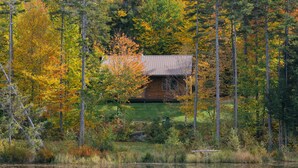 Eine Hütte, umgeben von einem Wald mit Herbstlaub, ein Teich im Vordergrund und eine Hügelkette in der Ferne.