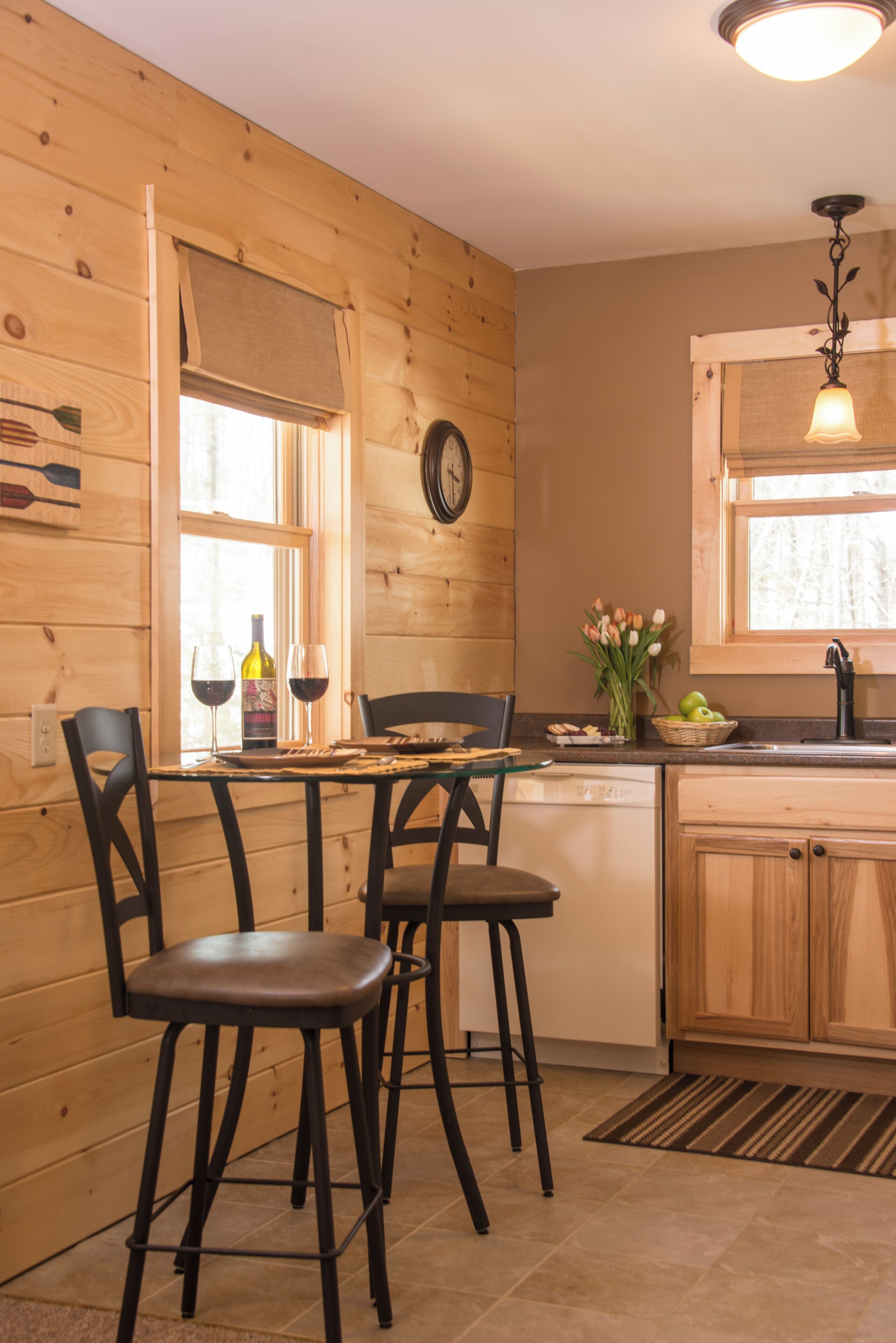 A wooden interior with a dining table, chairs, a clock, a window with blinds, and a kitchen area with a sink and countertop.