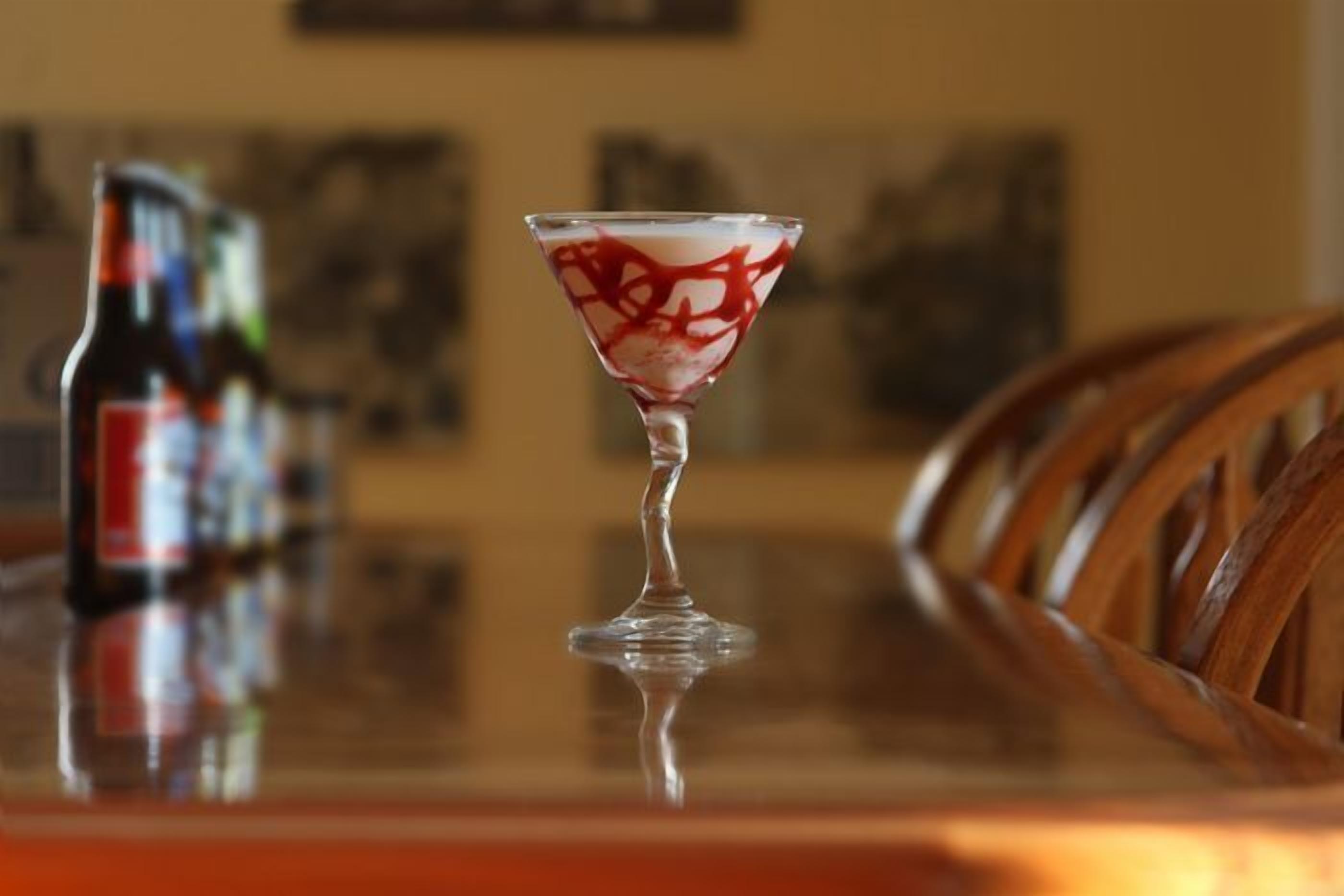 A martini glass with a red and white pattern on a reflective surface, a bottle of beer, and wooden chairs in the background.