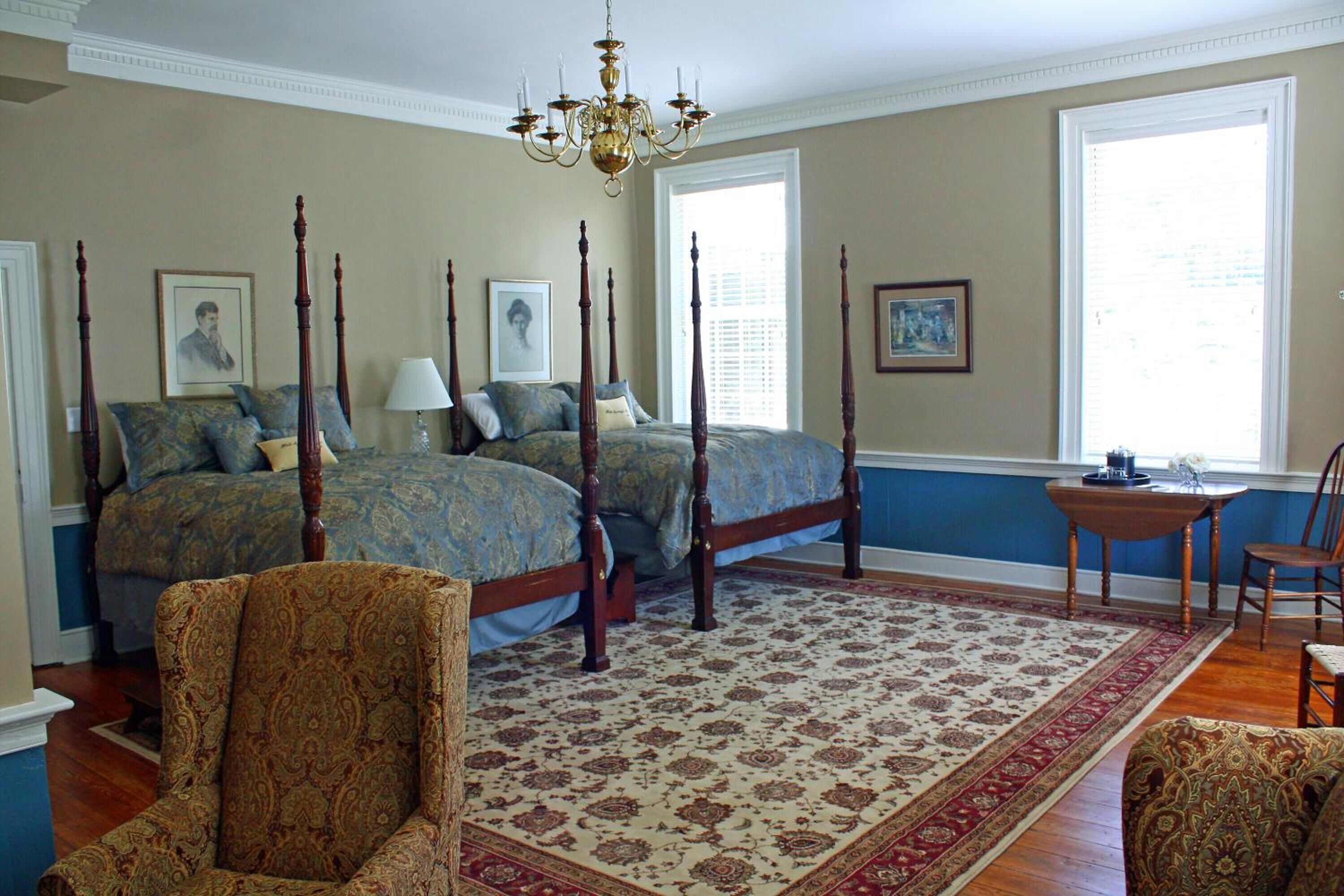 A bedroom with two four-poster beds, a chandelier, a large rug, and a view of the outdoors through a window.