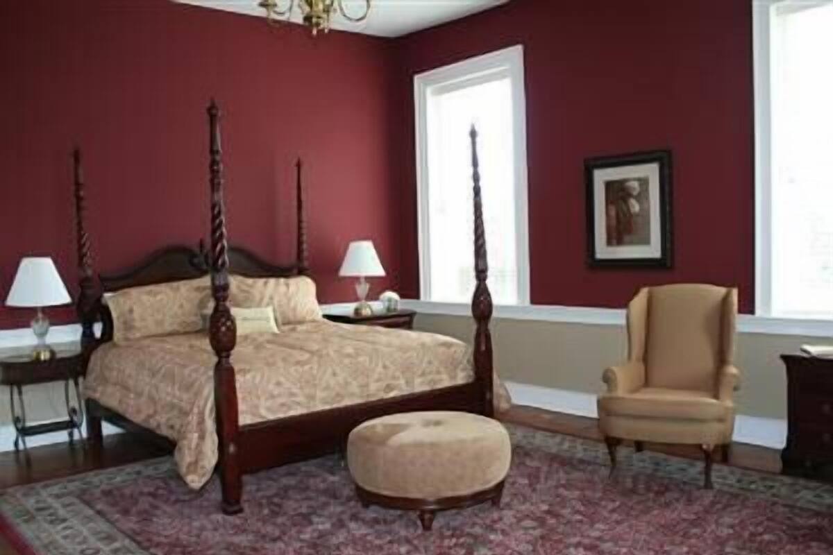 A bedroom with a four-poster bed, a side table, a chair, and a large window.