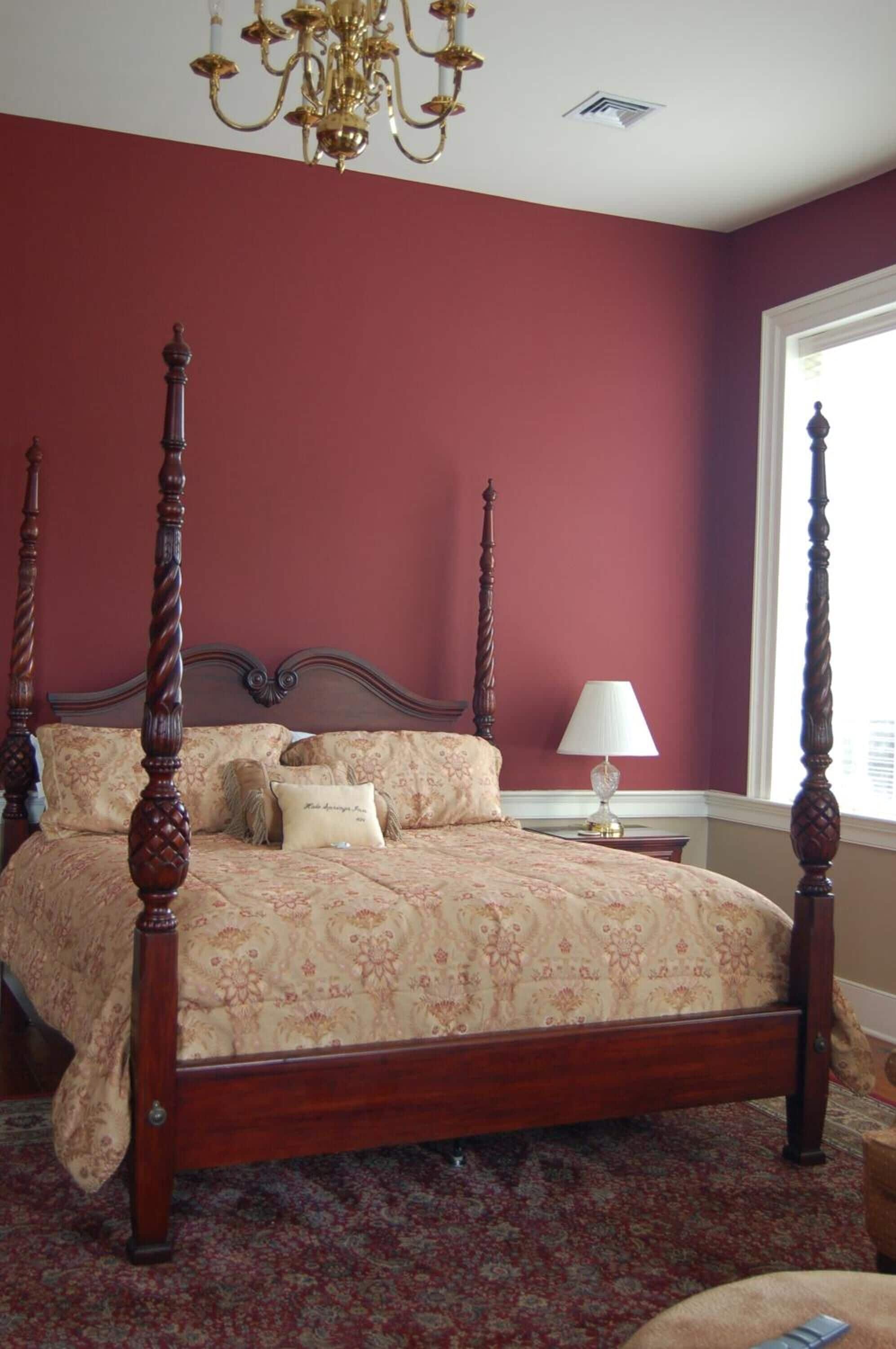 A four-poster bed with a floral bedspread and two pillows, a chandelier, and a nightstand with a lamp.
