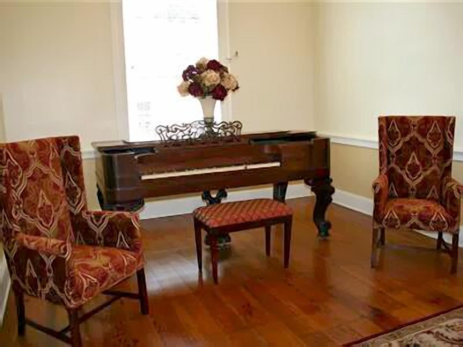 Una habitación con un piano de cola, dos sillones estampados y un florero sobre piso de madera.