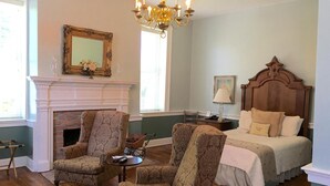 A room with a fireplace, two armchairs, a bed, a chandelier, and a mirror on the wall.