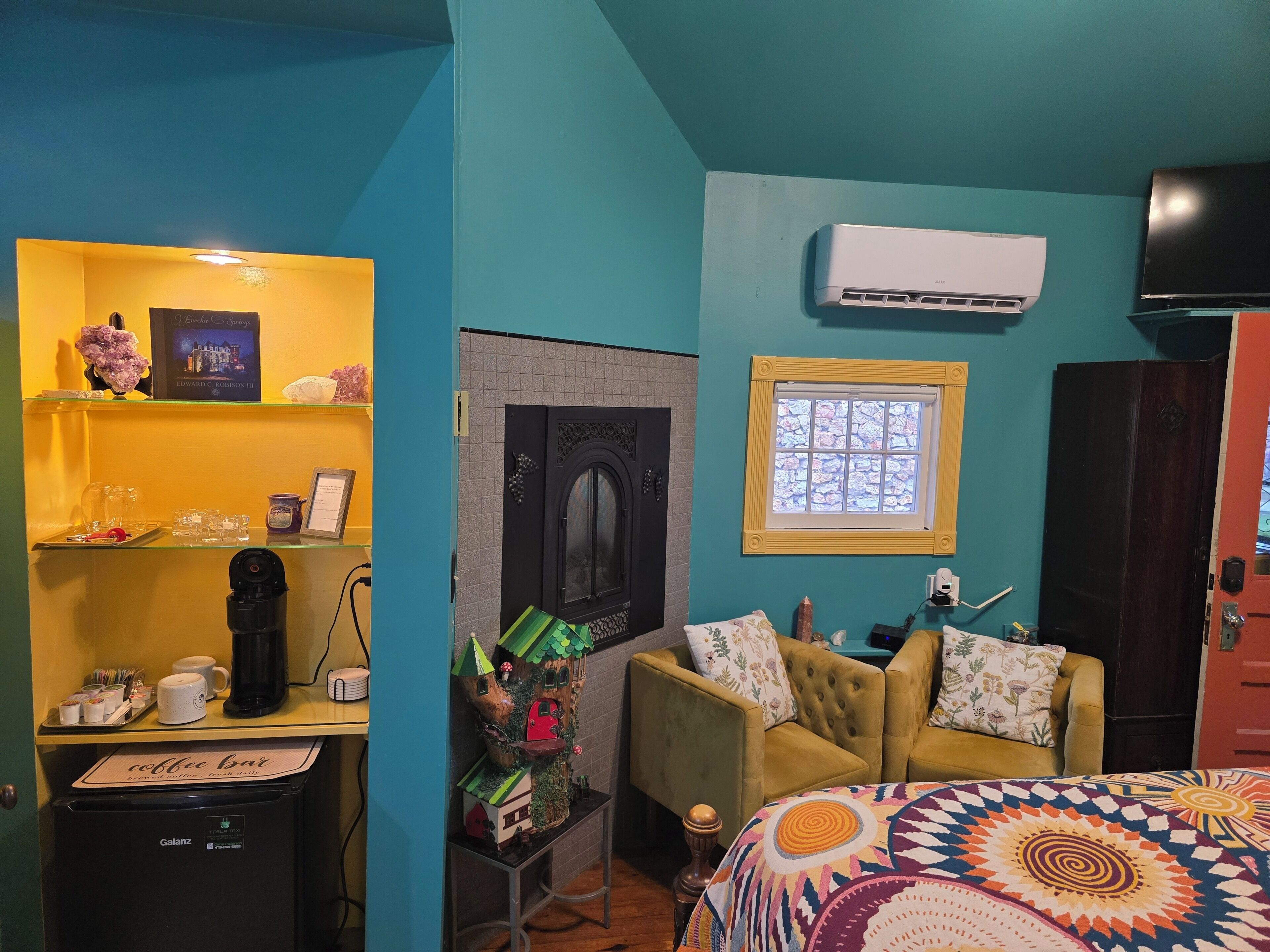 A room with a coffee bar, a small treehouse decoration, a bed with a colorful blanket, a mounted air conditioner, and a window with a view.