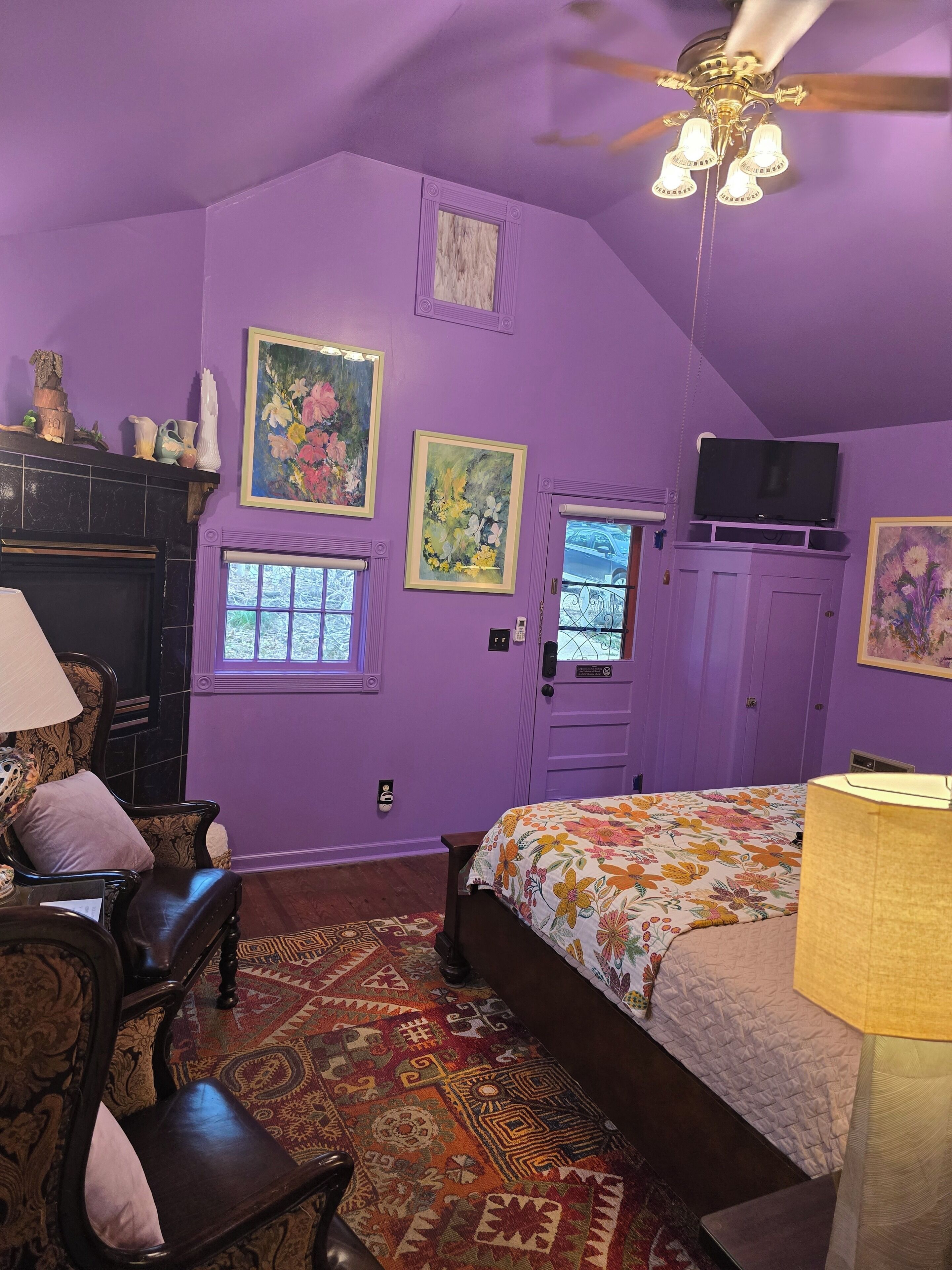 Un dormitorio con decoración morada, que incluye una cama, un escritorio, una silla, un televisor y un ventilador de techo.