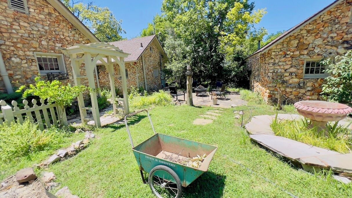 A stone building with a covered patio, a wheelbarrow, and a garden area.