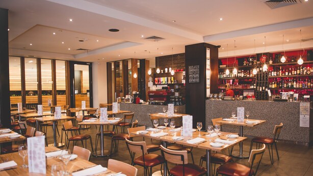A restaurant with multiple tables and chairs, a bar area with shelves of bottles, and a menu stand.