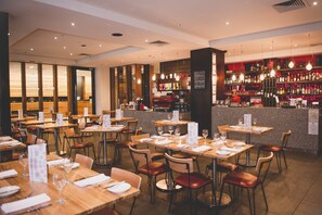 A restaurant with multiple tables and chairs, a bar area with shelves of bottles, and a menu stand.