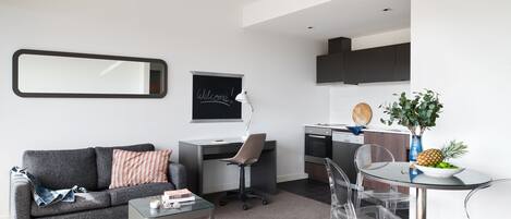 Una sala moderna con un sofá gris, una mesa de centro de vidrio, un escritorio con una silla, y una zona de cocina con una mesa de comedor y sillas.