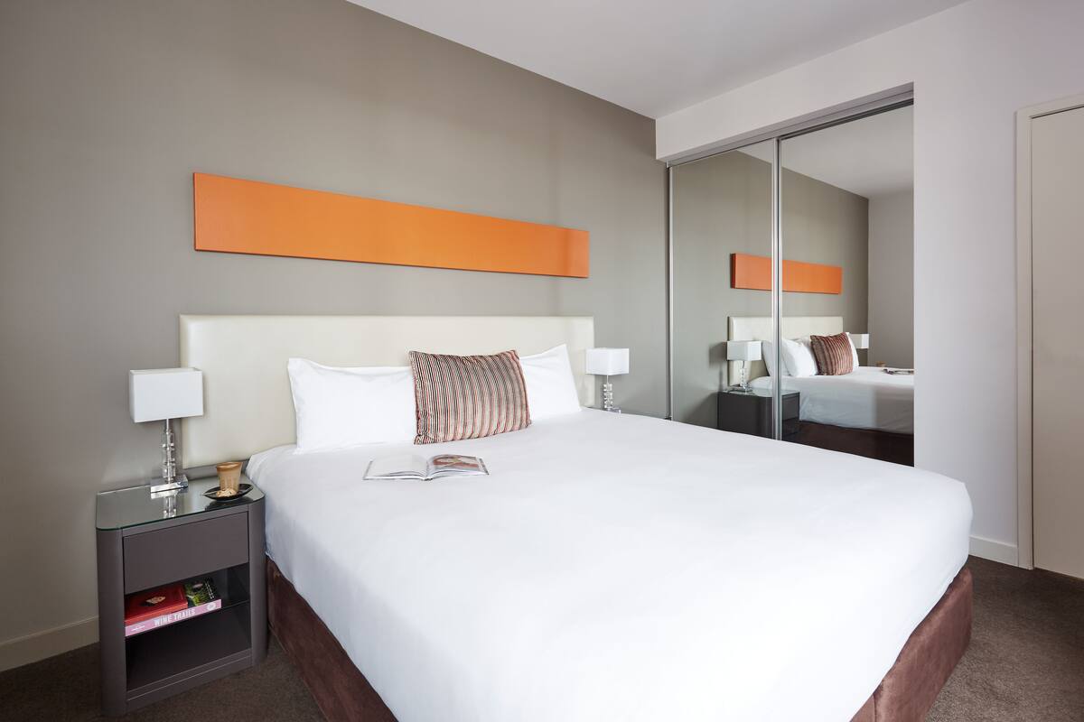 A hotel room with a large bed, a nightstand with a lamp, a bookshelf, and a mirror on the wall.
