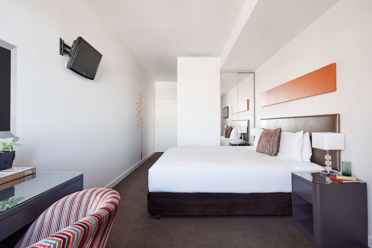 A modern hotel room with a large bed, a mounted TV, a bedside table with a lamp, and a striped chair.
