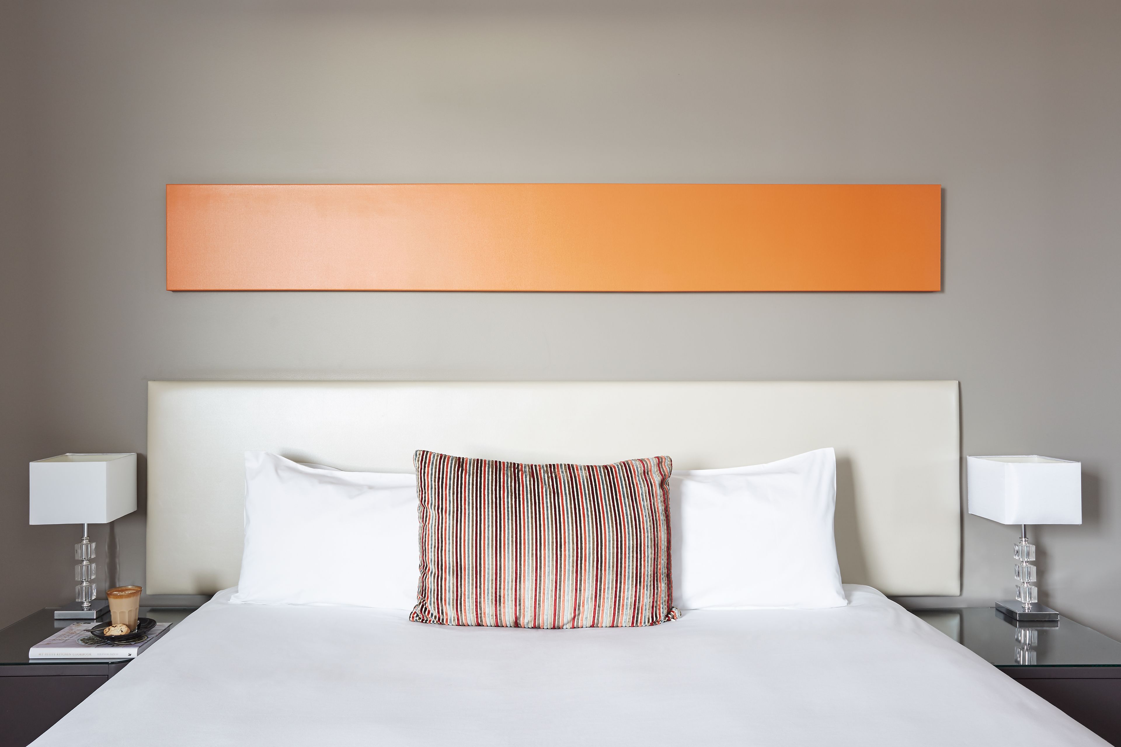 Et hotelværelse med en stor seng, to sengeborde med lamper, et orange kunstværk på væggen og en stribet pude.