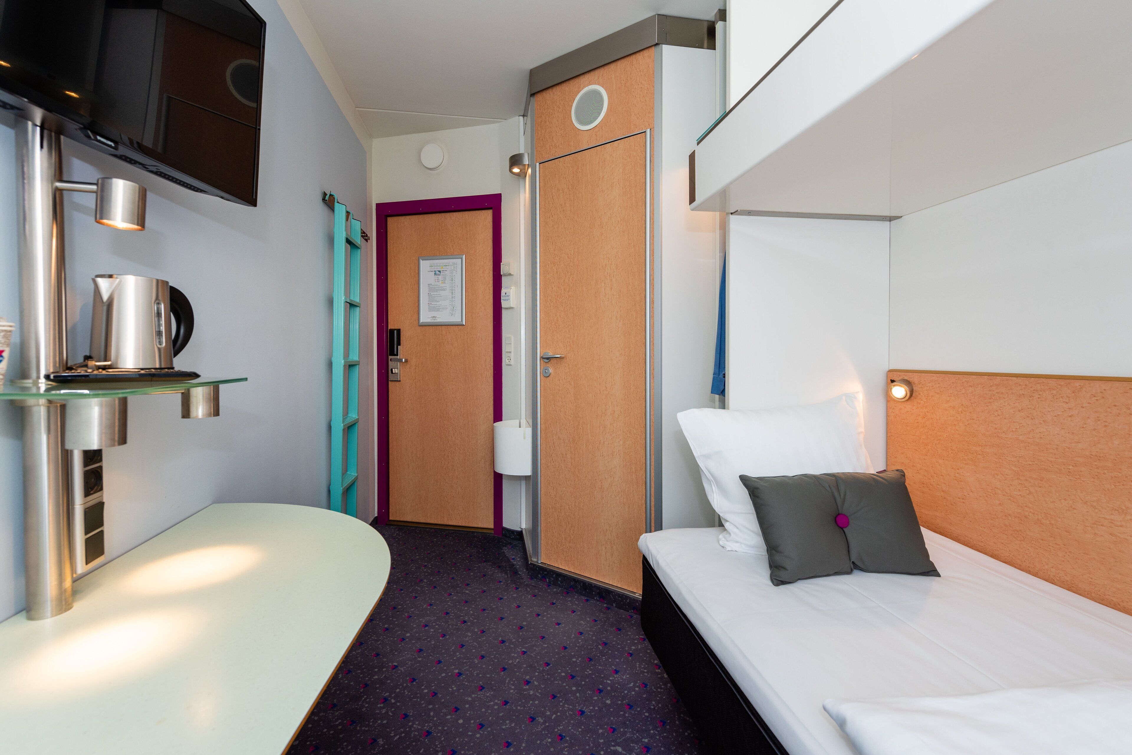 A modern hotel room with a bed, a small table with a kettle, a TV mounted on the wall, and a door with a purple frame.