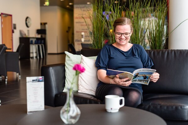 A person sitting on a black leather couch reading a magazine, with a white mug and a vase with a pink flower on a table in front of them.