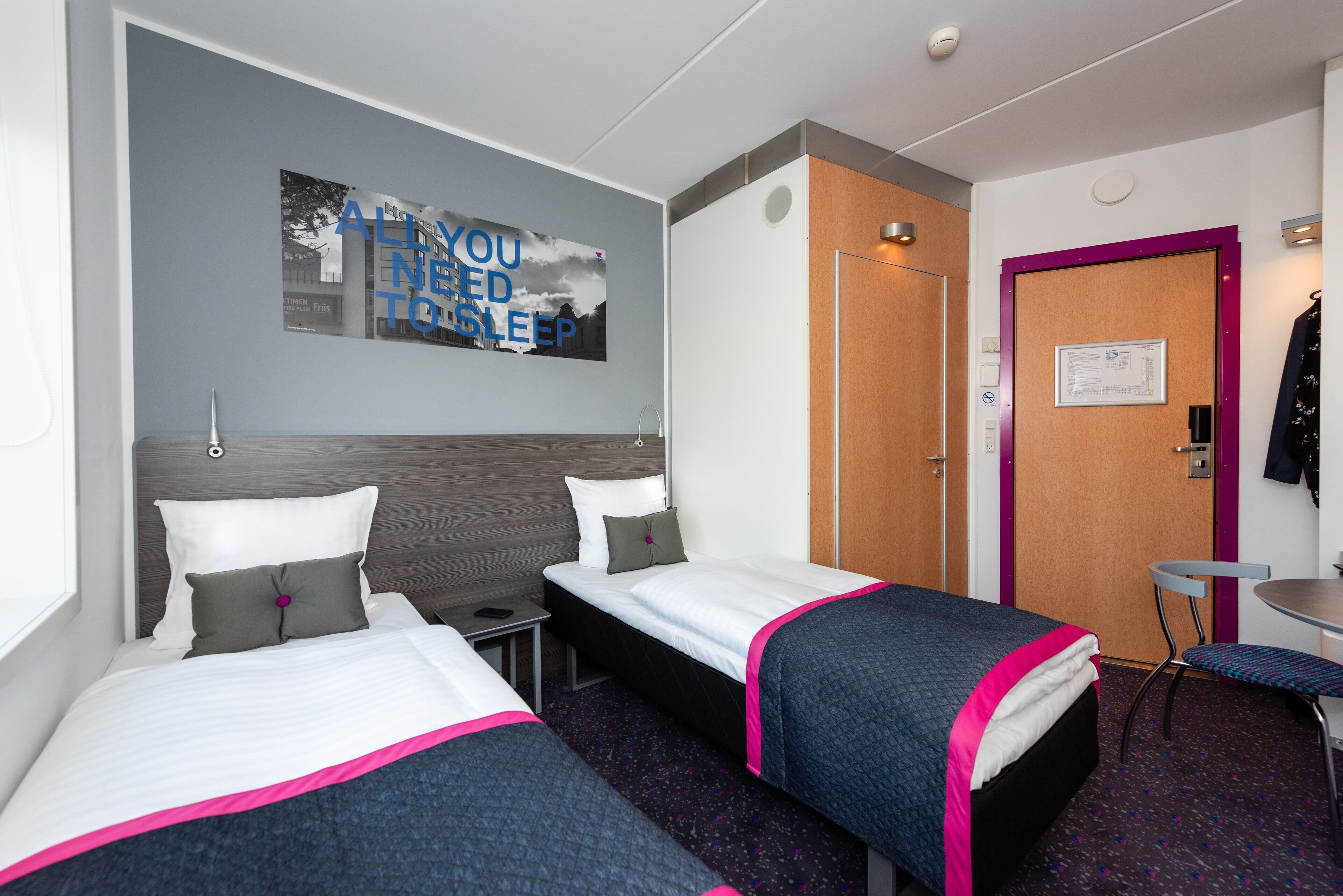 Habitación de hotel con dos camas, una mesita, una silla, una puerta y un cartel en la pared.