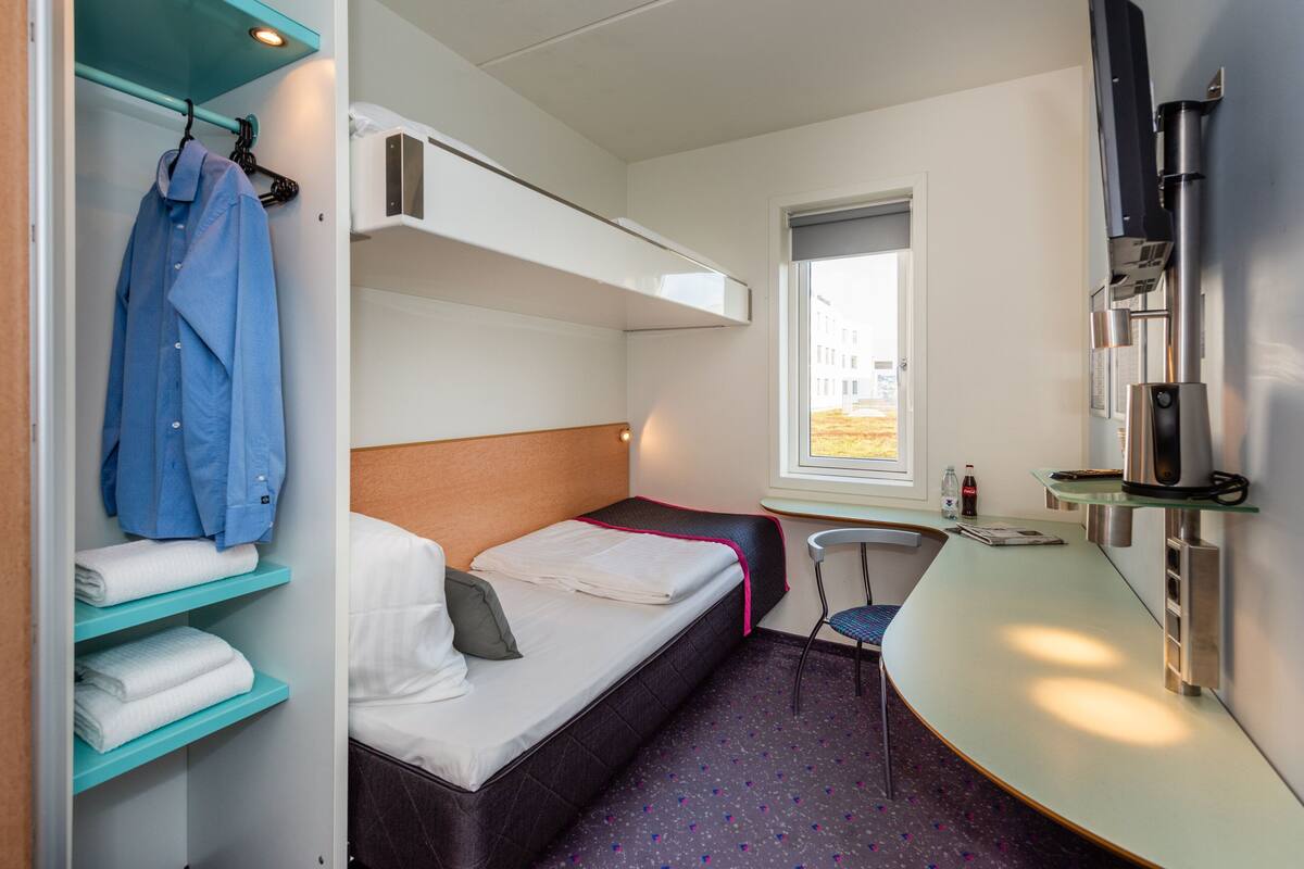 A compact hotel room with a bunk bed, a small desk with a chair, a window, and a TV mounted on the wall.