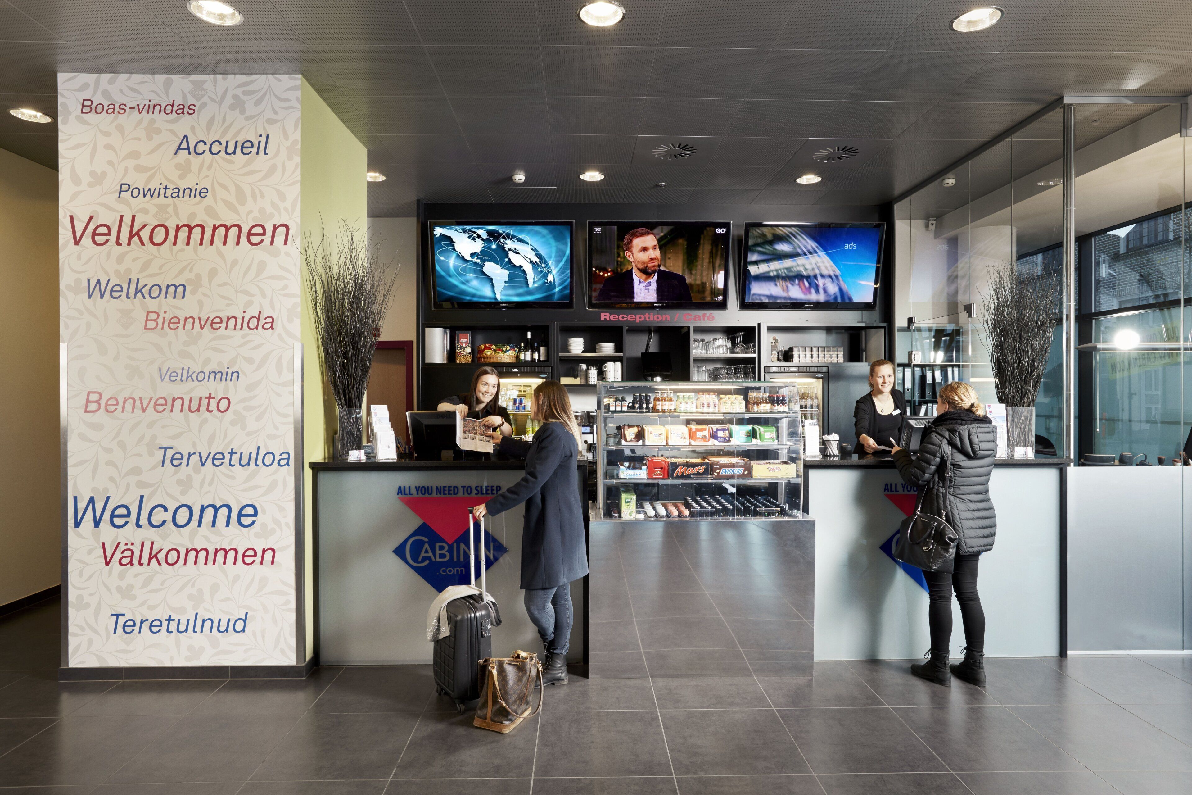 A modern hotel reception area with a large welcome sign displaying multiple languages, a reception desk, and a minibar.
