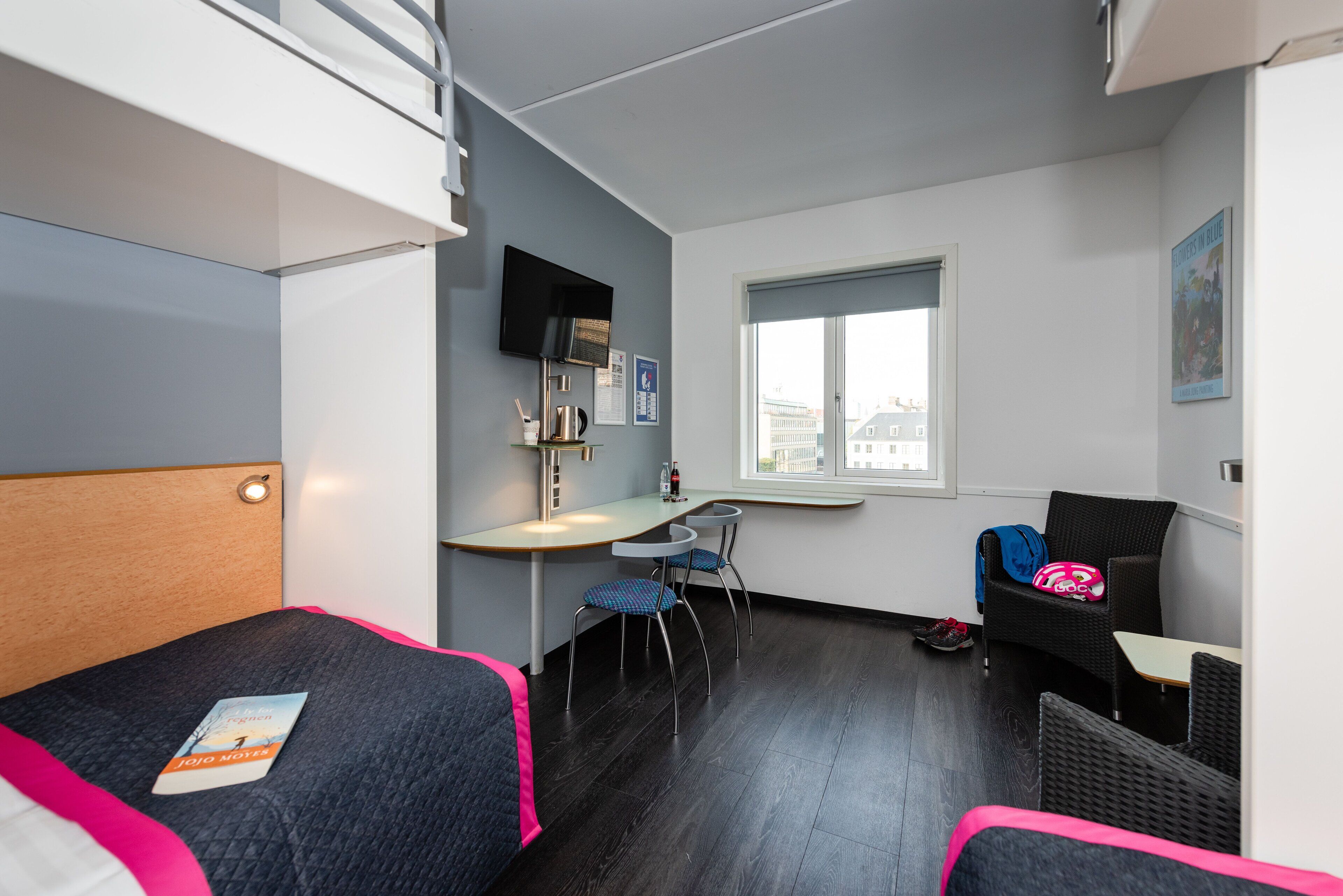 Ein kleines Hotelzimmer mit einem Bett, einem Schreibtisch, zwei Stühlen, einem Fenster und einem an der Wand montierten Fernseher.